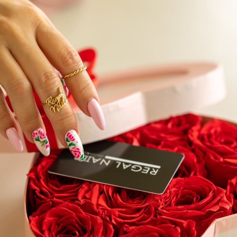 Uñas almendradas rosa pastel con rosas pintadas a mano, junto a una caja de rosas rojas y la tarjeta Regal Nails